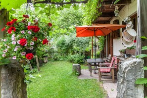 Une terrasse avec une table et des chaises, entourée de verdure et agrémentée d’un parasol rouge suspendu.