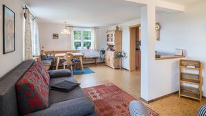 A living room with a sofa, a dining table with chairs, a kitchen area with cabinets, and a window with plants.
