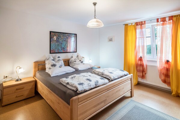 A bedroom with a wooden bed, a nightstand, a desk lamp, a framed artwork, and a window with curtains.
