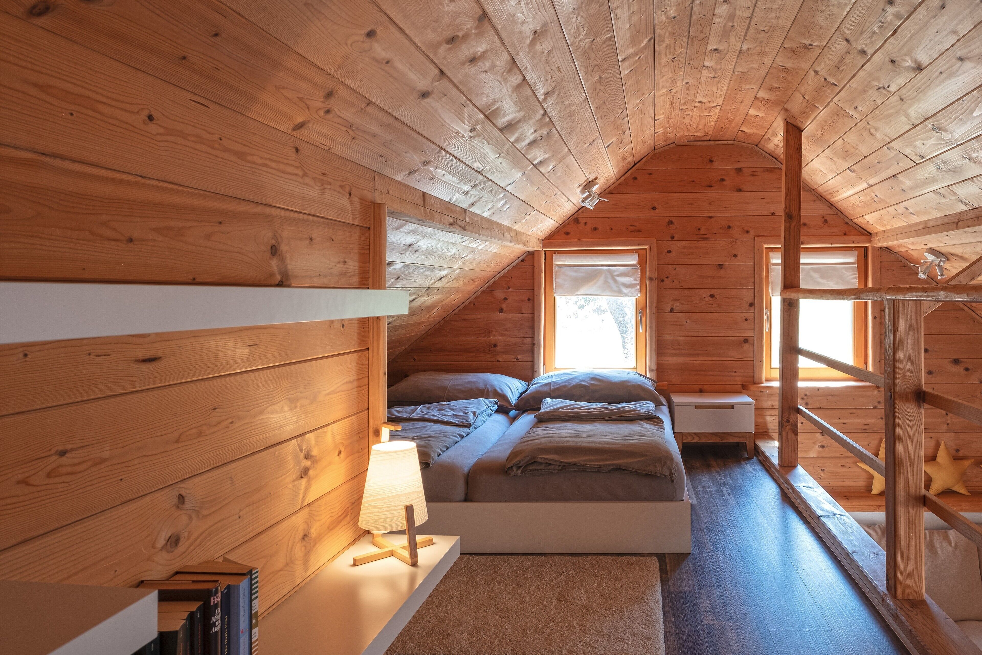 Ein gemütliches Schlafzimmer im Dachgeschoss mit schrägverlegtem Holzboden, einem Bett mit Kissen, einem Nachttisch mit Lampe und einem Fenster mit Blick.