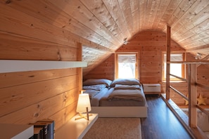 A cozy attic bedroom with a wooden sloped ceiling, a bed with pillows, a bedside table with a lamp, and a window with a view.