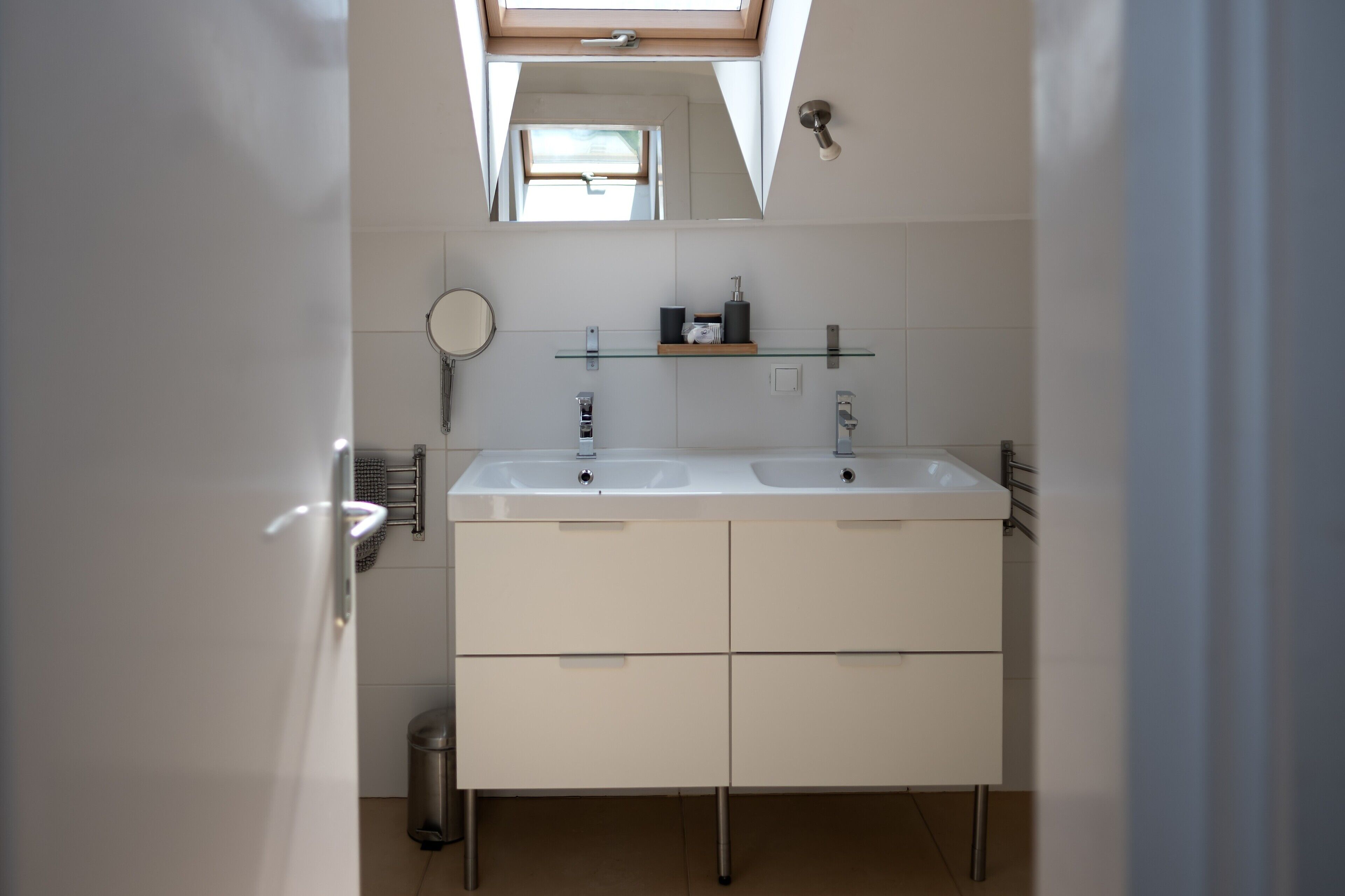 A modern bathroom with a skylight, two sinks, a mirror, and a glass shelf with toiletries.