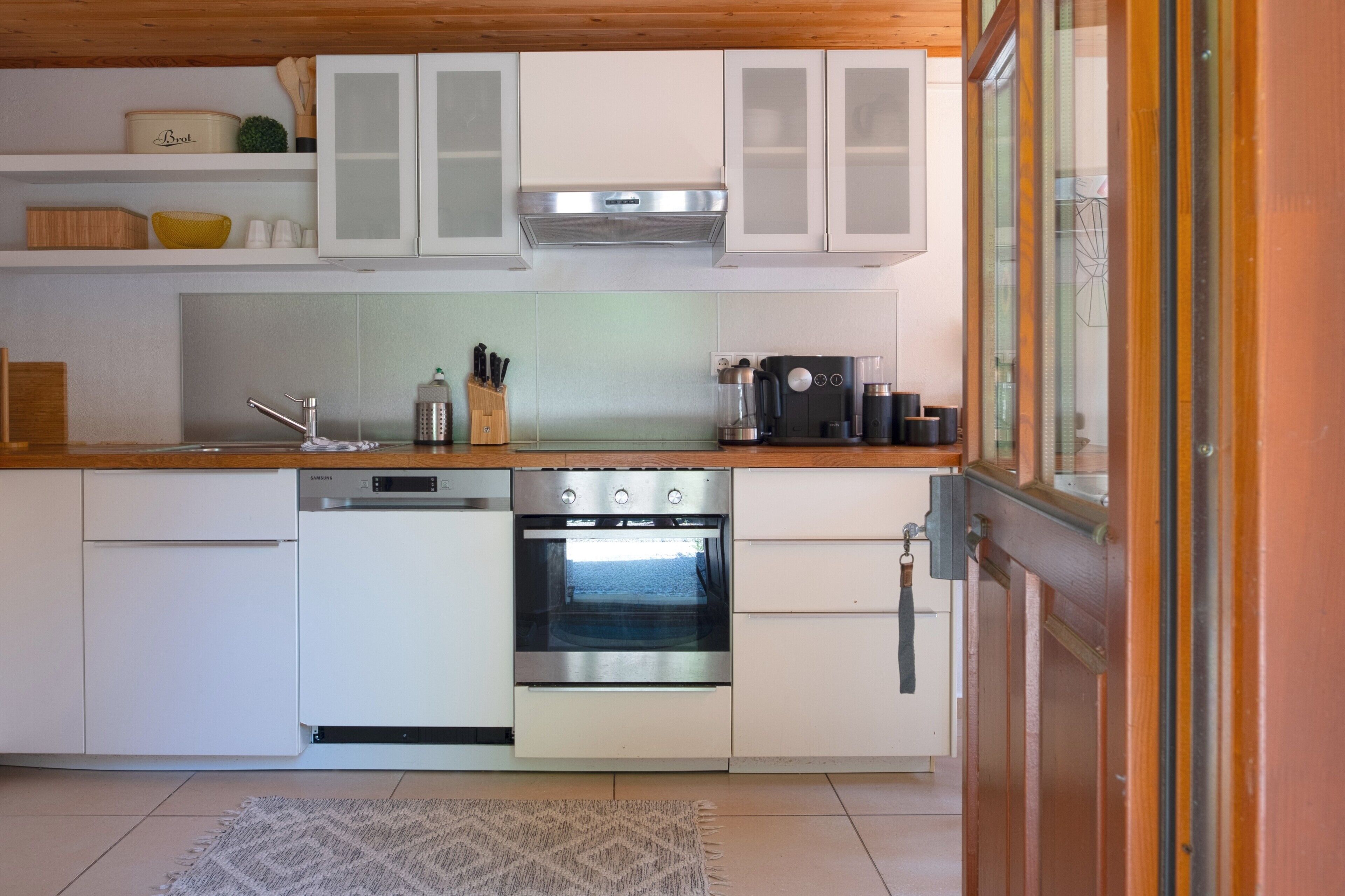 Una cocina compacta con muebles blancos, horno de acero inoxidable, lavavajillas y cafetera.