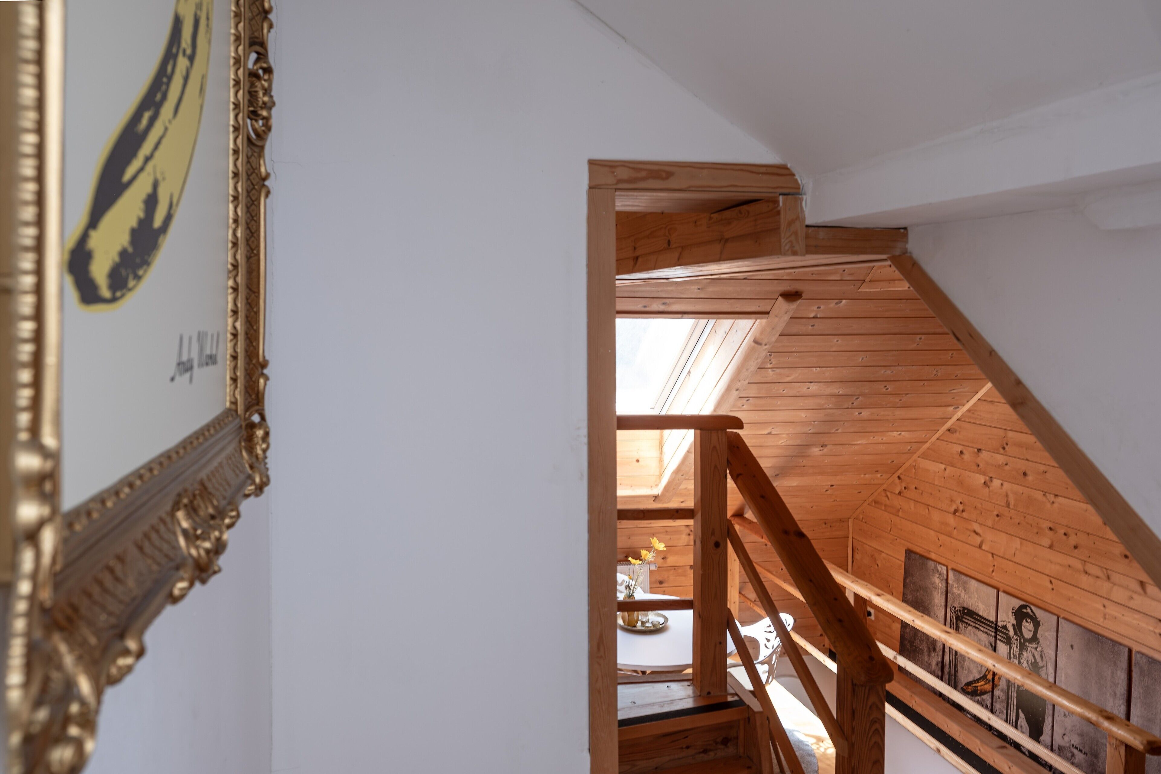 A wooden staircase leading to an upper floor with a skylight, a framed picture on the wall, and a table with a vase of flowers.