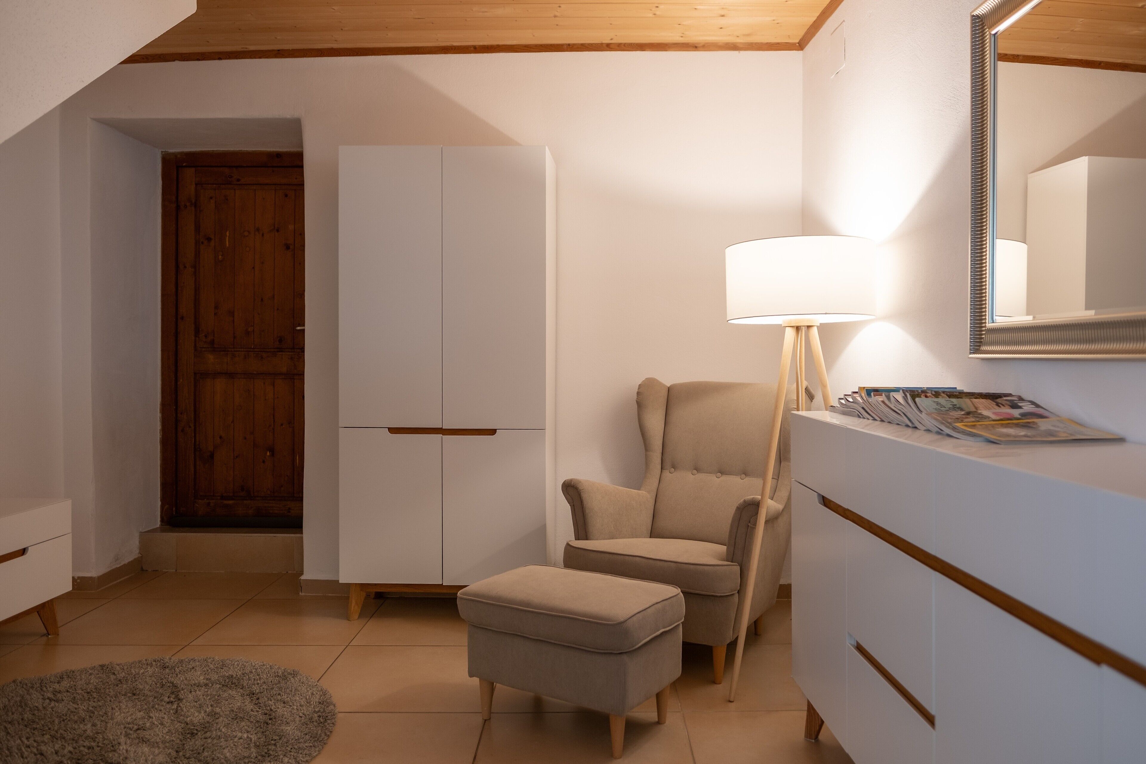 A room with a wooden door, a white wardrobe, a beige armchair, a small ottoman, a floor lamp, and a magazine on a dresser.