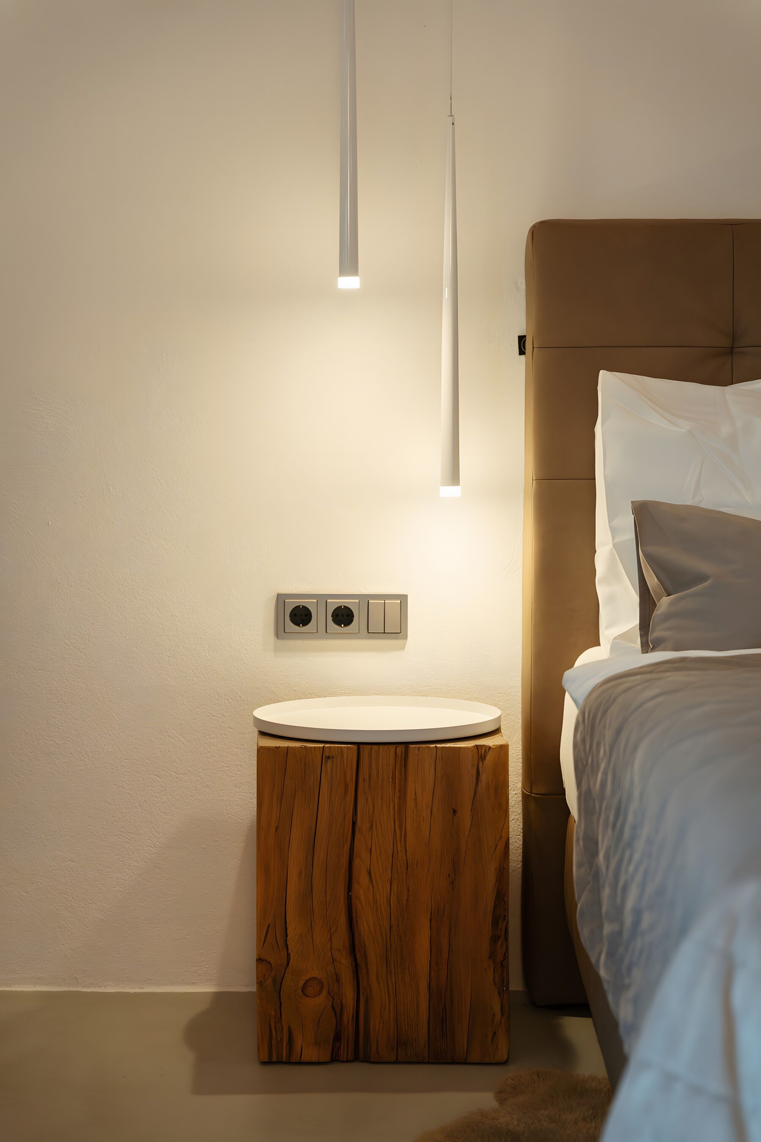 A modern bedroom with a round wooden nightstand, a bed with a headboard, and a wall-mounted light fixture.