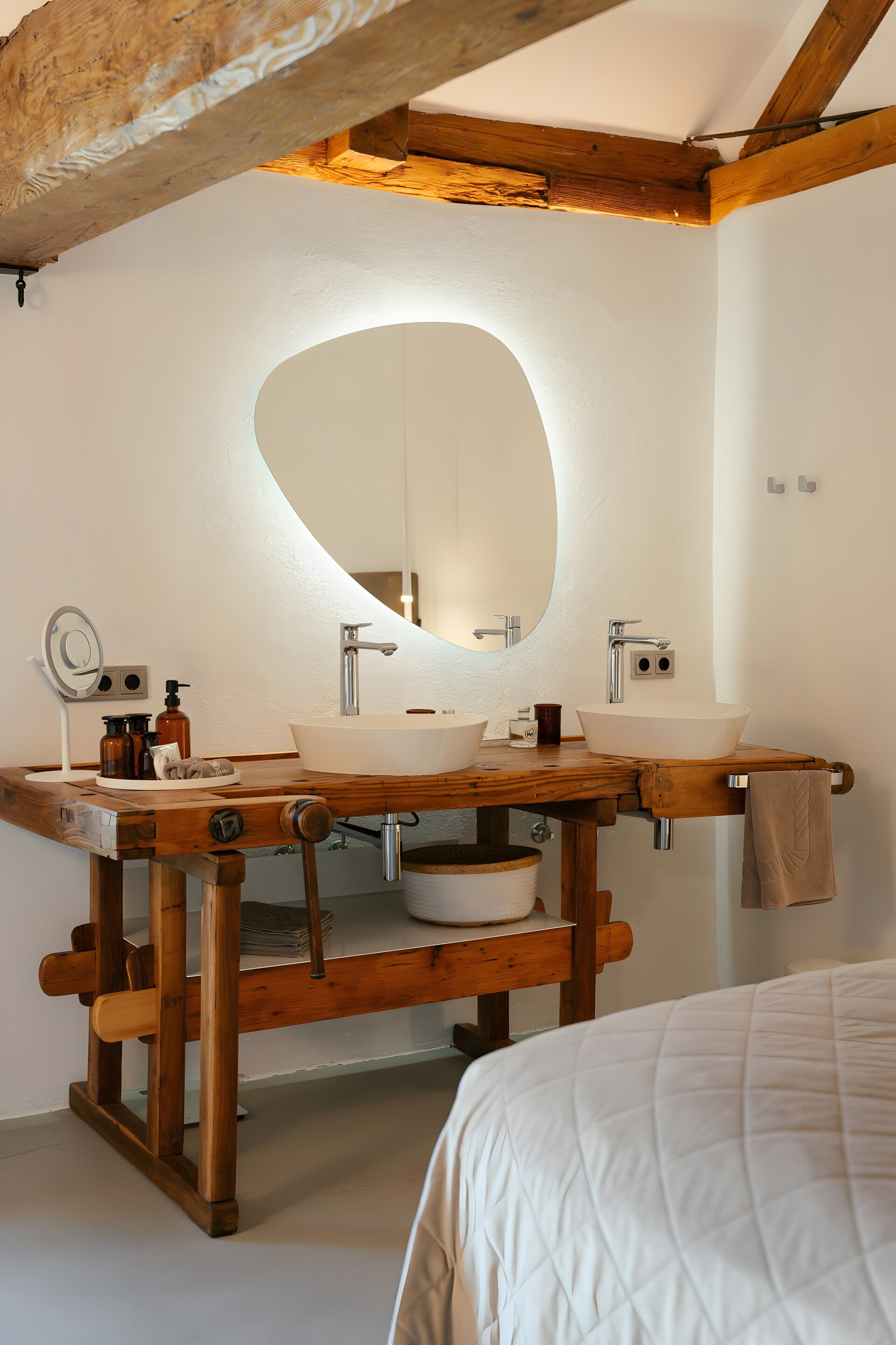 A bathroom with a wooden vanity, two sinks, a large mirror, and a bed in the foreground.