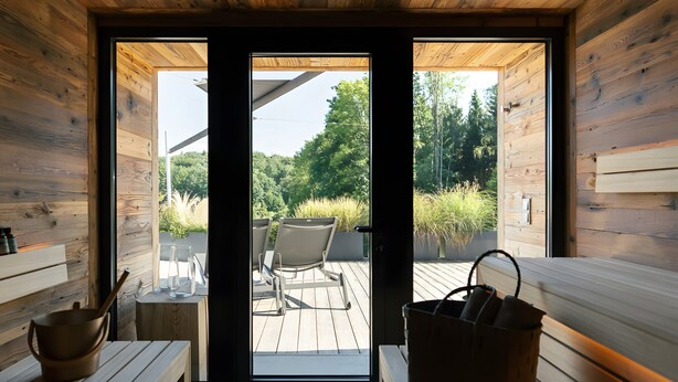 Een sauna met houten banken, een bankje met een mand, en uitzicht op een terras buiten met stoelen en planten.