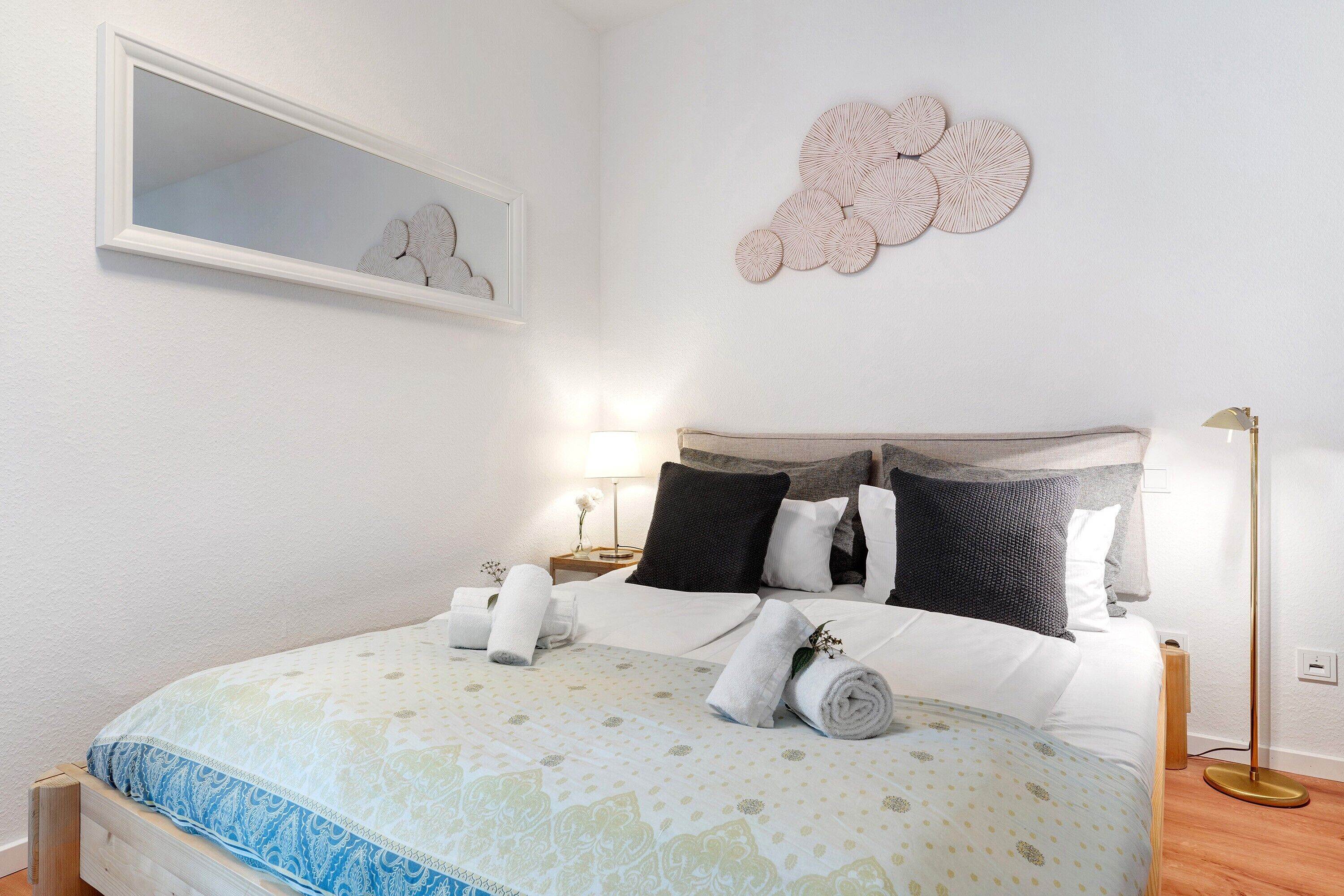 Una cama bien hecha, toallas enrolladas, una mesita de noche con lámpara y un adorno en la pared.