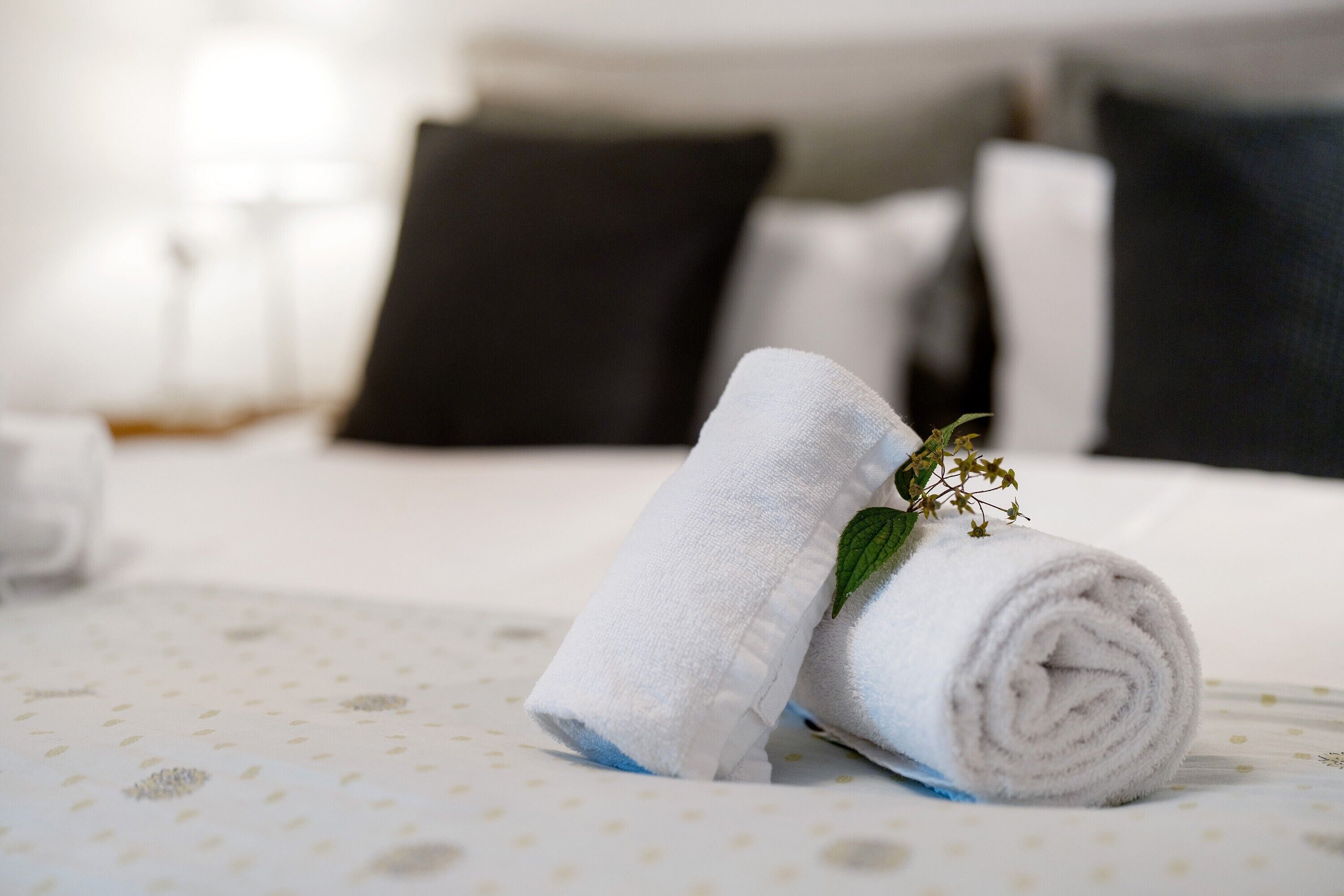 Een keurig opgevouwen witte handdoek met een groene blad, geplaatst op een bed met stippen en zwarte kussens op de achtergrond.