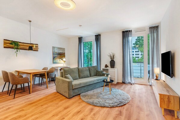 Un salon moderne comprenant un canapé, une table à manger, des chaises, un téléviseur et une grande fenêtre avec des rideaux.