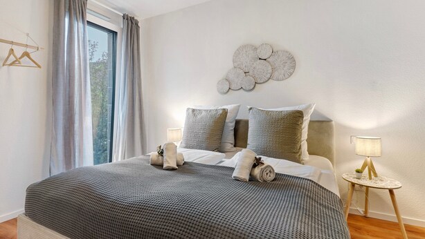 A bedroom with a large bed, a bedside table with a lamp, a wall decoration, and a window with curtains.