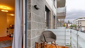 A balcony with a wicker chair, a wooden stool, and a view of a cityscape.