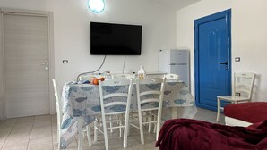 A small, clean room with a dining table, chairs, a TV, a refrigerator, and a blue door.