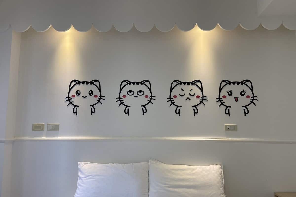 Un dormitori amb un llit, roba de llit blanca i quatre vinils murals de gatets de dibuix animat.