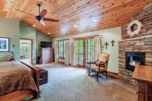 A spacious bedroom with a wooden ceiling, a fireplace, and a large window.