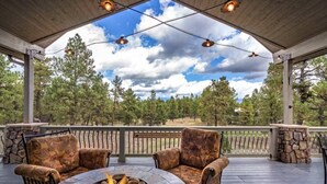 En udendørs terrasse med en ildsted i sten, to møbler med armlæn i et mønstret design, og udsigt til et skovområde under et overdækket område med hængende lys.