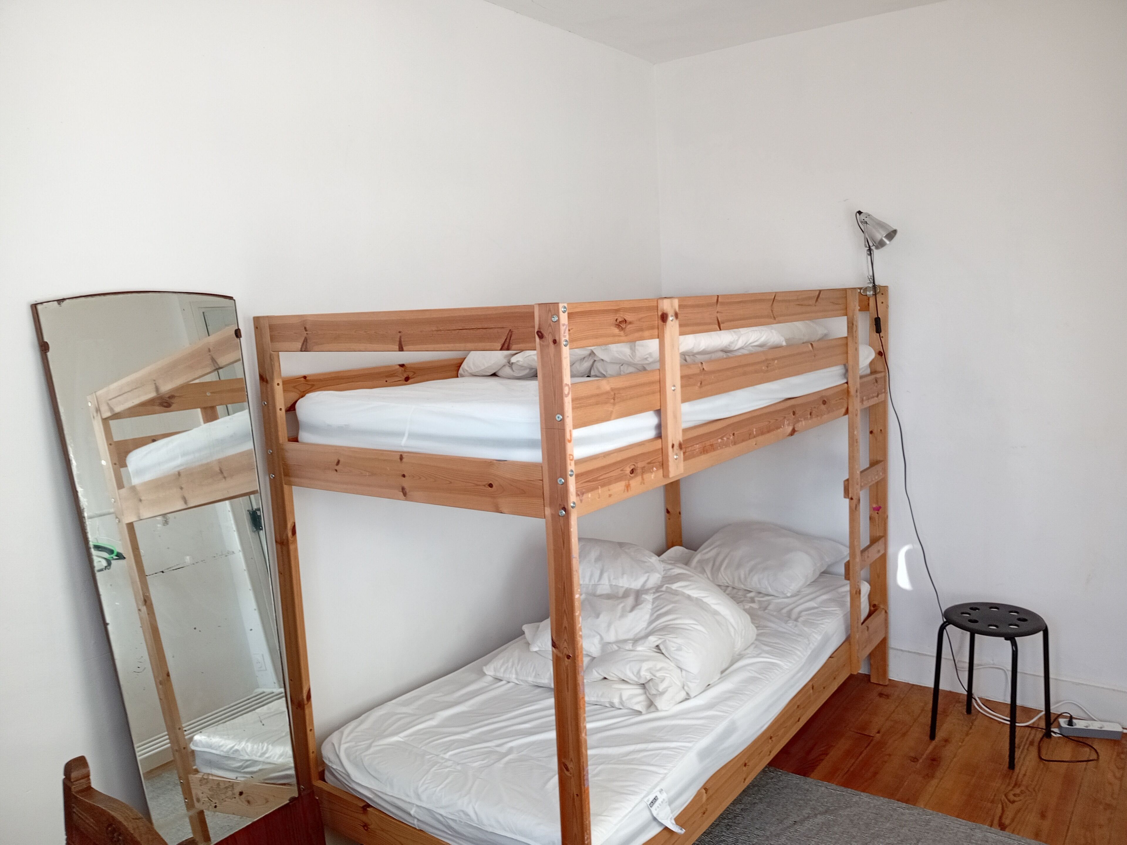 A bunk bed with a wooden frame, white bedding, a mirror, a desk lamp, and a power outlet.