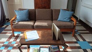 Un salon avec un canapé marron, deux coussins bleus, un meuble en bois orné d’un tableau encadré et une petite table sur laquelle se trouvent des papiers et des livres.
