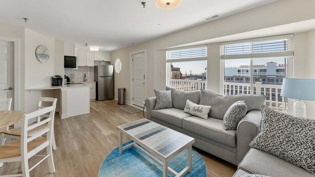 A modern apartment with a kitchenette, a dining area, a living room with a sofa and a coffee table, and large windows offering a city view. - 2BR Condo with Modern Amenities Steps to Beach! (Wildwood)