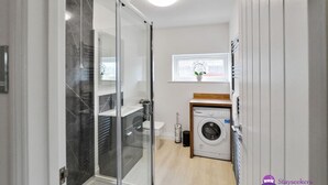A modern bathroom with a glass shower enclosure, a built-in washing machine, and a small window with a plant.