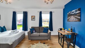 A modern room with a bed, a grey sofa, a dining table with chairs, and a blue wall with a painting.