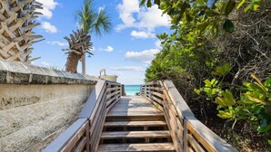 Un escalier en bois menant à une plage, entouré de verdure et sous un ciel dégagé.