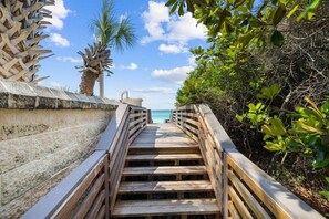 A wooden staircase leading to a beach, surrounded by greenery and a clear sky.