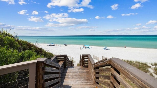 Un escalier en bois mène à une plage où l’eau est d’un bleu turquoise limpide, et où quelques parasols de plage sont installés.