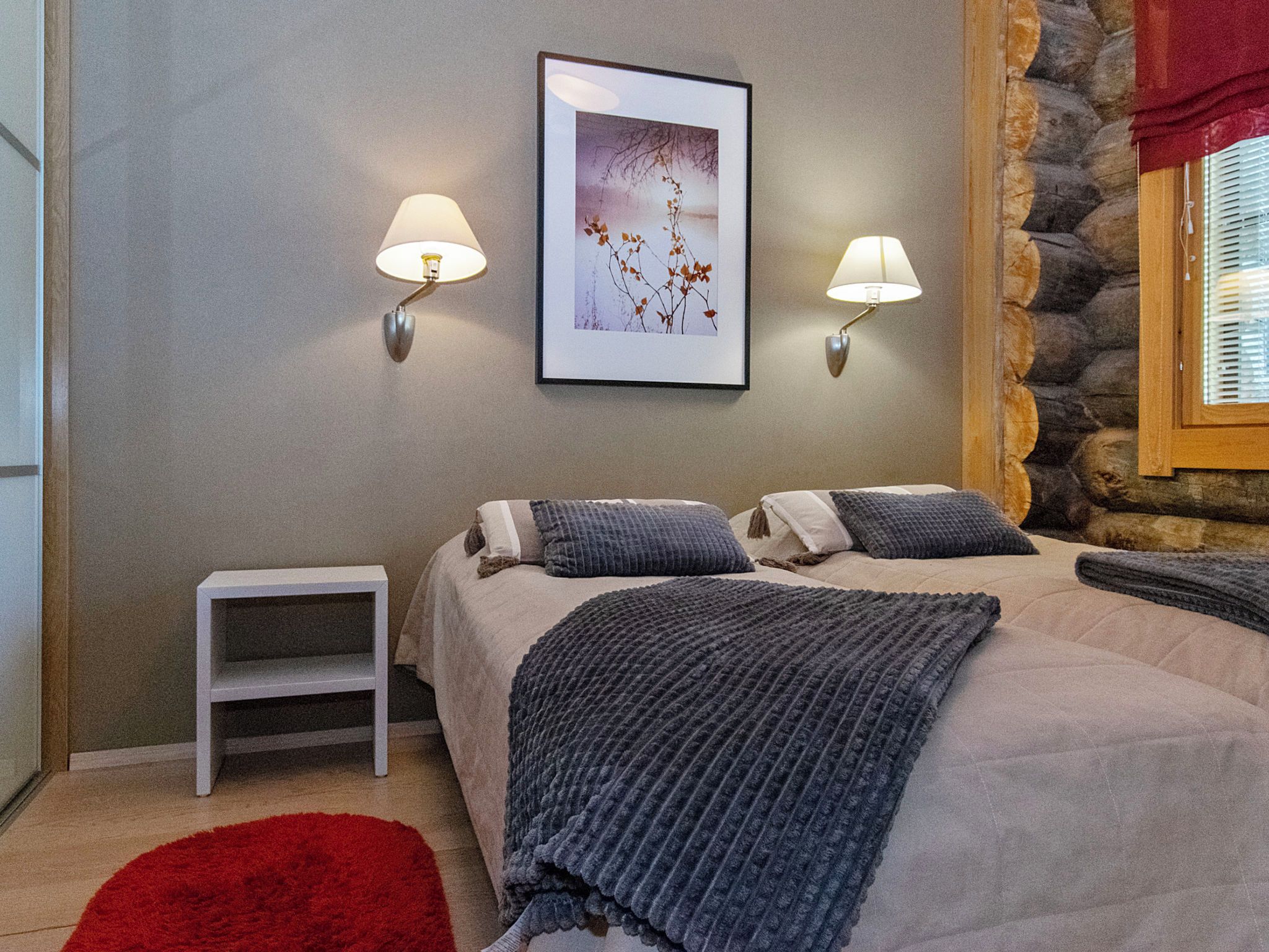 A bedroom with a bed, two bedside lamps, a small table, a red rug, and a framed picture on the wall.