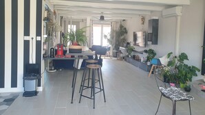 A modern living room with a kitchen area, featuring a dining table, bar stools, and potted plants. - Villa Nyvioni, 3 ch, spa et Piscine (Agde)