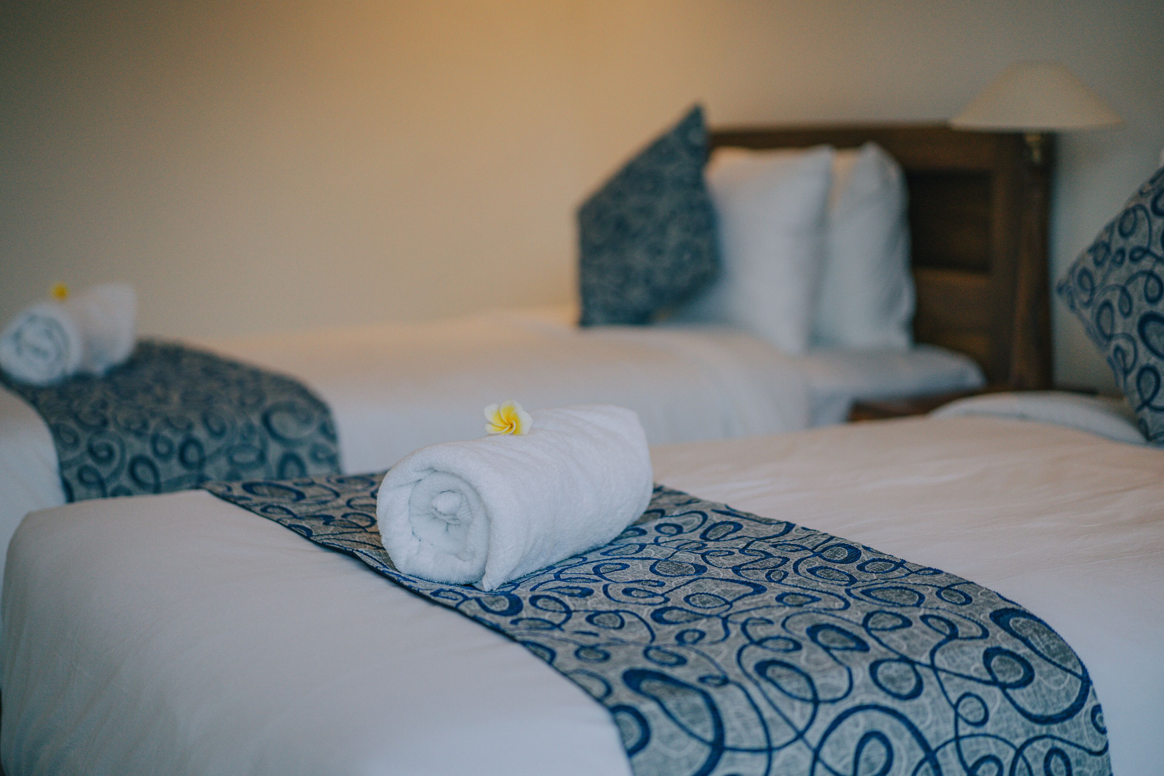 Ein Hotelzimmer mit zwei Betten, jedes mit einem aufgerollten weißen Handtuch und einem blauen Kissen mit Muster.