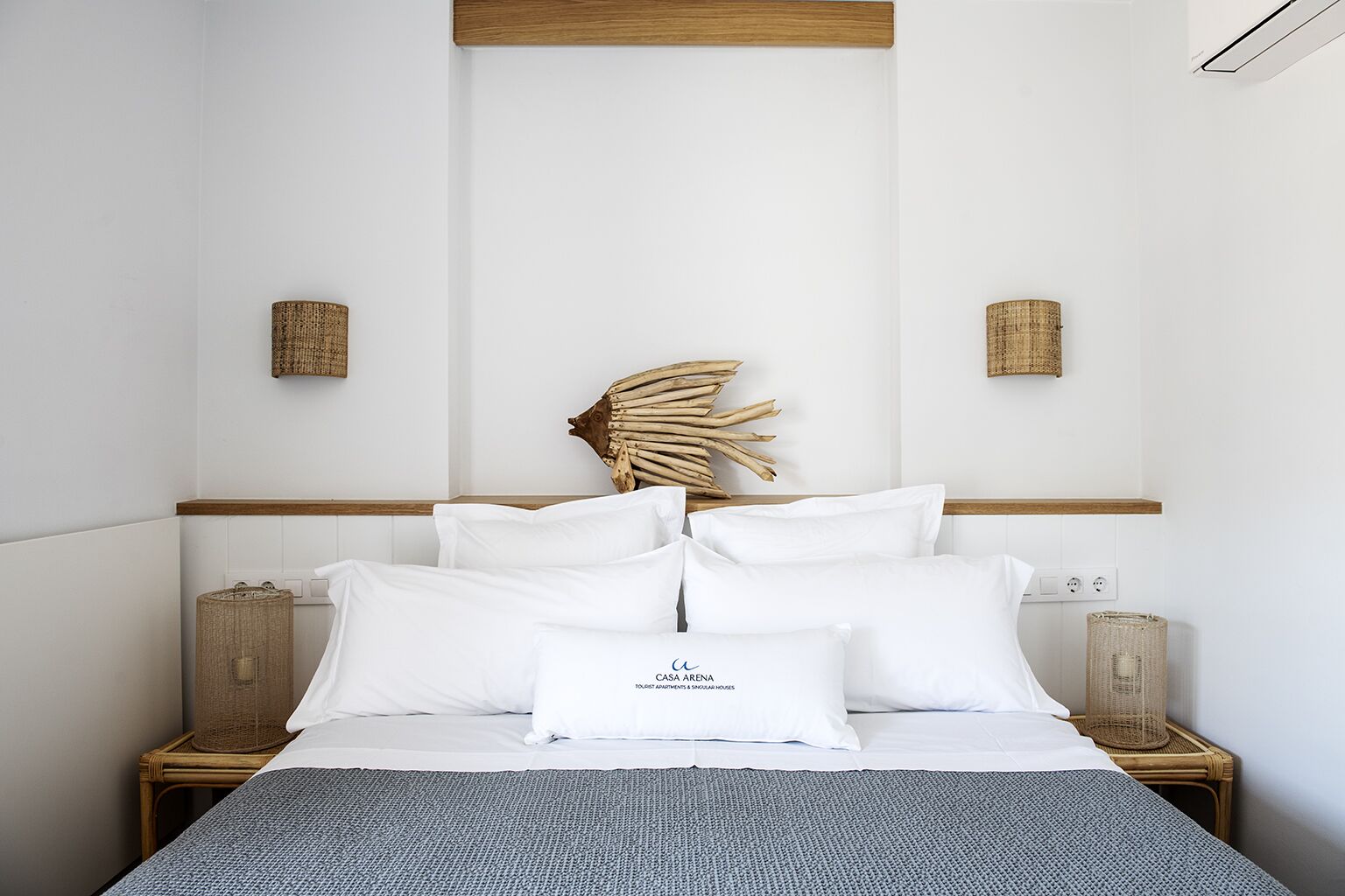 Een slaapkamer met een bed, witte kussens, een decoratief hoofdbord, nachtkastjes en een geweven mand.
