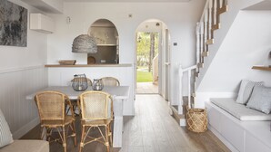 Una cocina moderna con armarios blancos, un comedor con sillas de mimbre y una escalera que sube a un dormitorio estilo loft.
