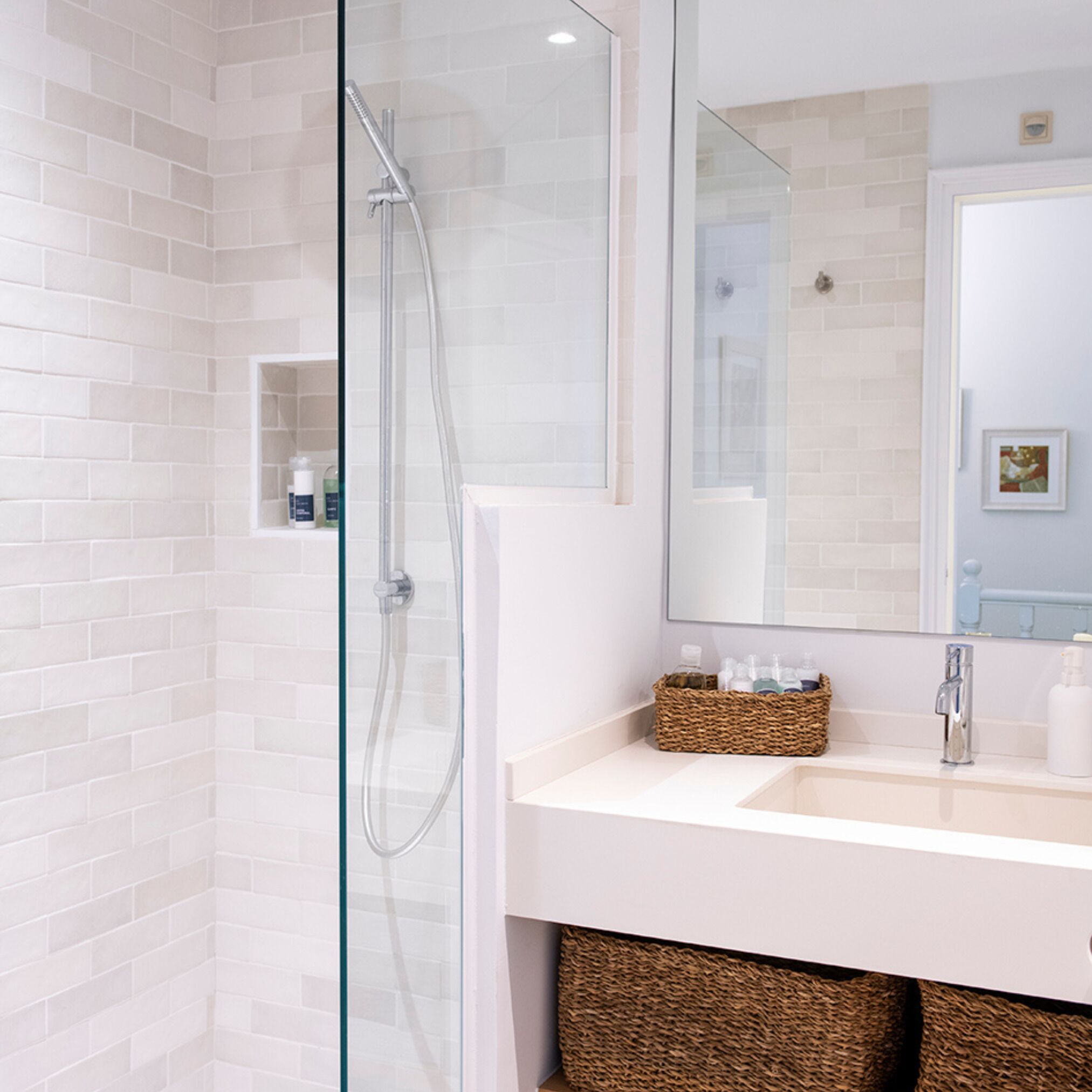 Un baño moderno con ducha envuelta en vidrio, paredes de azulejos blancos, un espejo grande y un lavamanos blanco con una cesta de mimbre.
