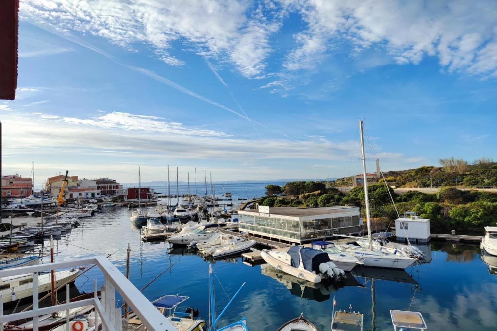 A marina with multiple boats docked, a building with a flat roof, and a clear sky with some clouds.