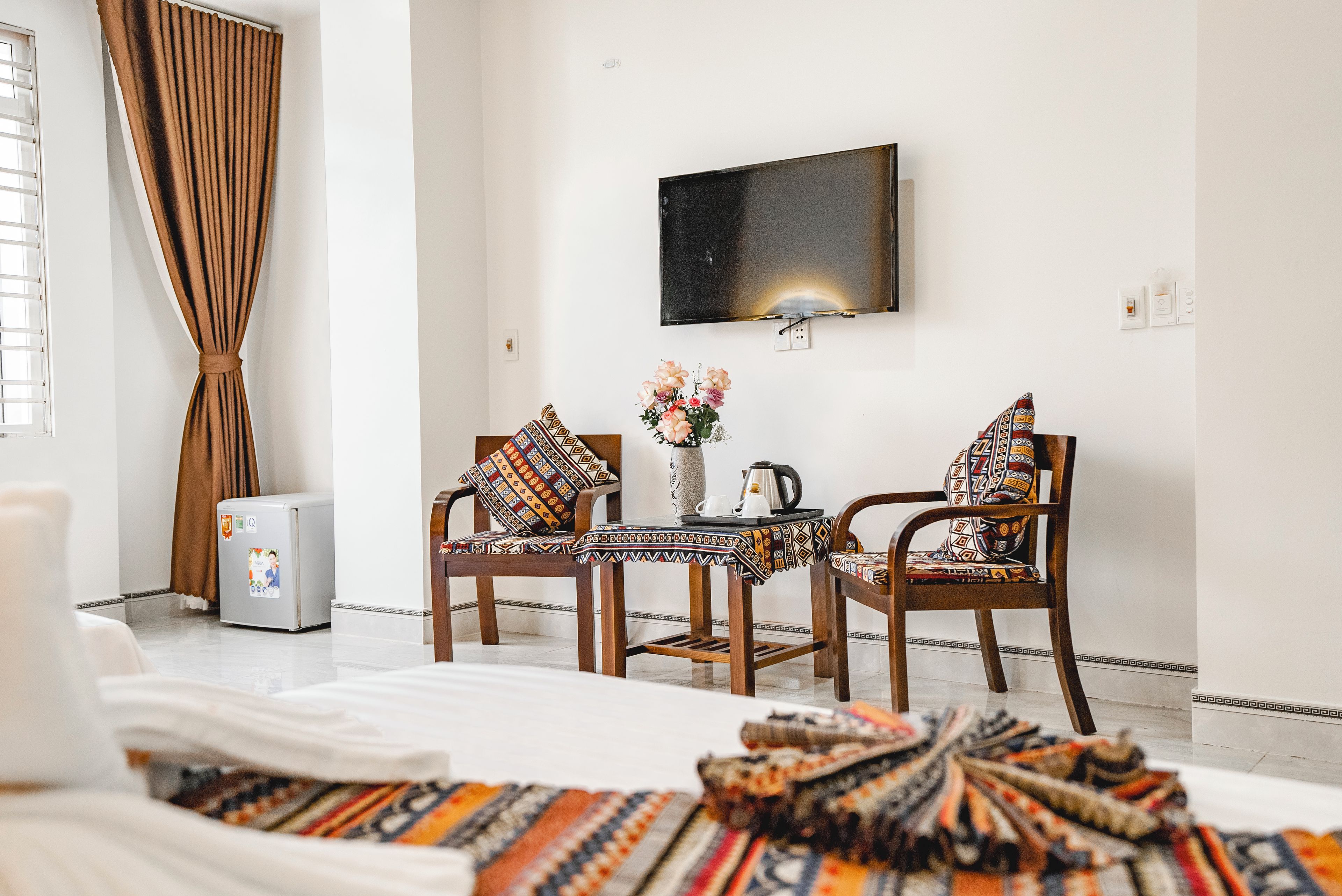 A hotel room with a bed, two chairs, a small table, a TV, and a window with curtains.