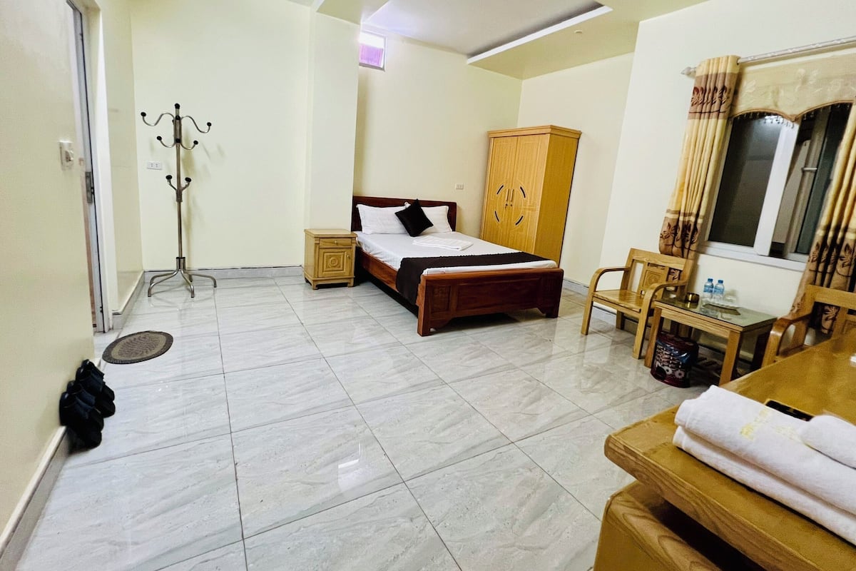 A hotel room with a bed, a wooden wardrobe, a desk with a chair, a coat rack, and a window with curtains.