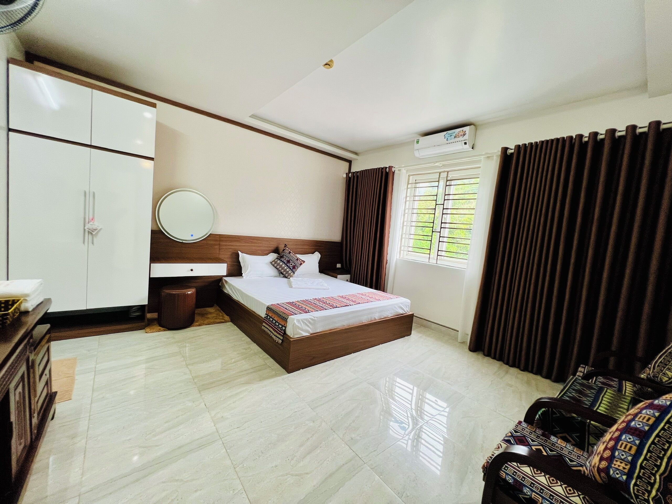 A hotel room with a bed, a bedside table, a wardrobe, a mirror, a window with curtains, and a ceiling fan.