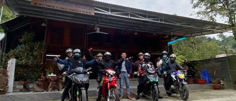 Un grupo de personas en motocicletas frente a un edificio con un letrero que dice 'Peaceful Mountain Homestay'.