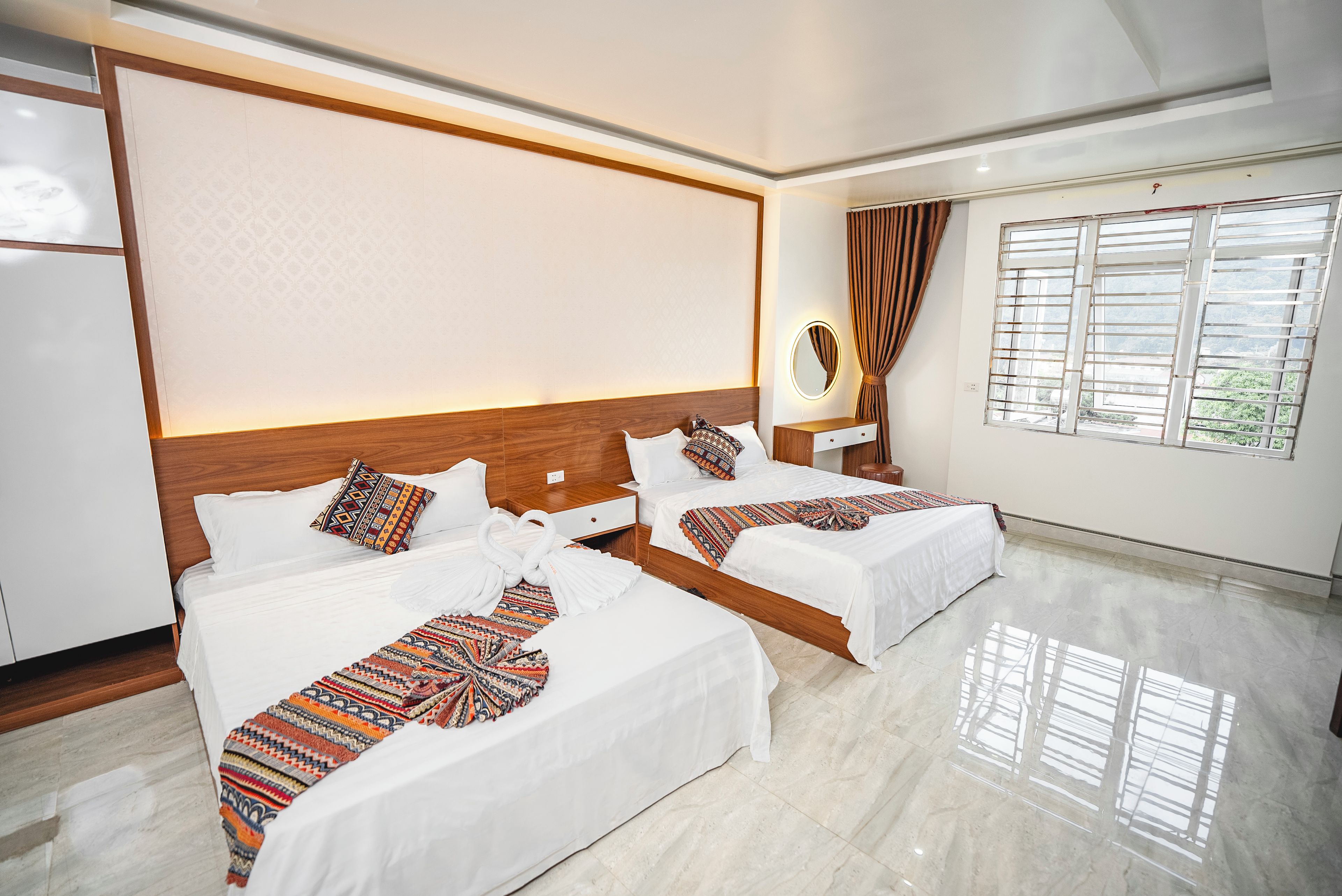 A hotel room with two beds, a wooden headboard, a bedside table with a mirror, and a window with curtains.