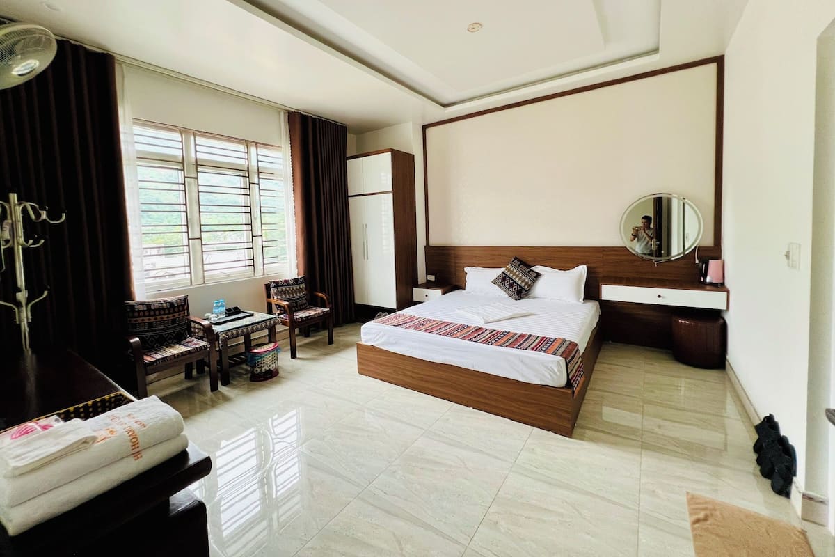 A hotel room with a large bed, a desk with a chair, a wardrobe, a mirror, and a window with curtains.