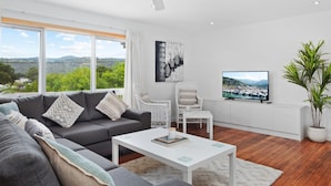 A modern living room with a grey sofa, white coffee table, and wicker chairs. A TV is mounted on the wall, and a large window offers a scenic view of greenery and hills. - House Allinga, great for large or multiple families. (Jindabyne)