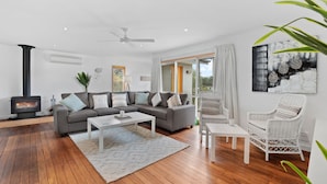 A modern living room with a grey sofa, white coffee table, and wicker chairs. A black wood-burning stove is visible in the corner. - House Allinga, great for large or multiple families. (Jindabyne)