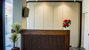 Un mostrador de recepción con diseño de listones de madera, una planta en maceta a la izquierda y un jarrón con flores rojas a la derecha.