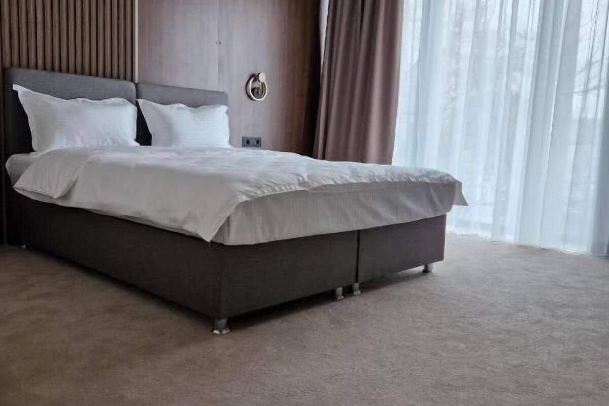 Une chambre d’hôtel moderne avec un grand lit, des murs lambrissés et une grande fenêtre ornée de rideaux légers.