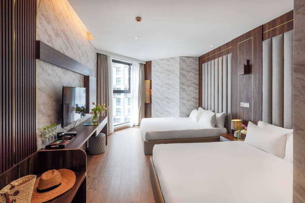 Habitación de hotel con dos camas, televisión, escritorio con computadora y ventana con vista a edificios.