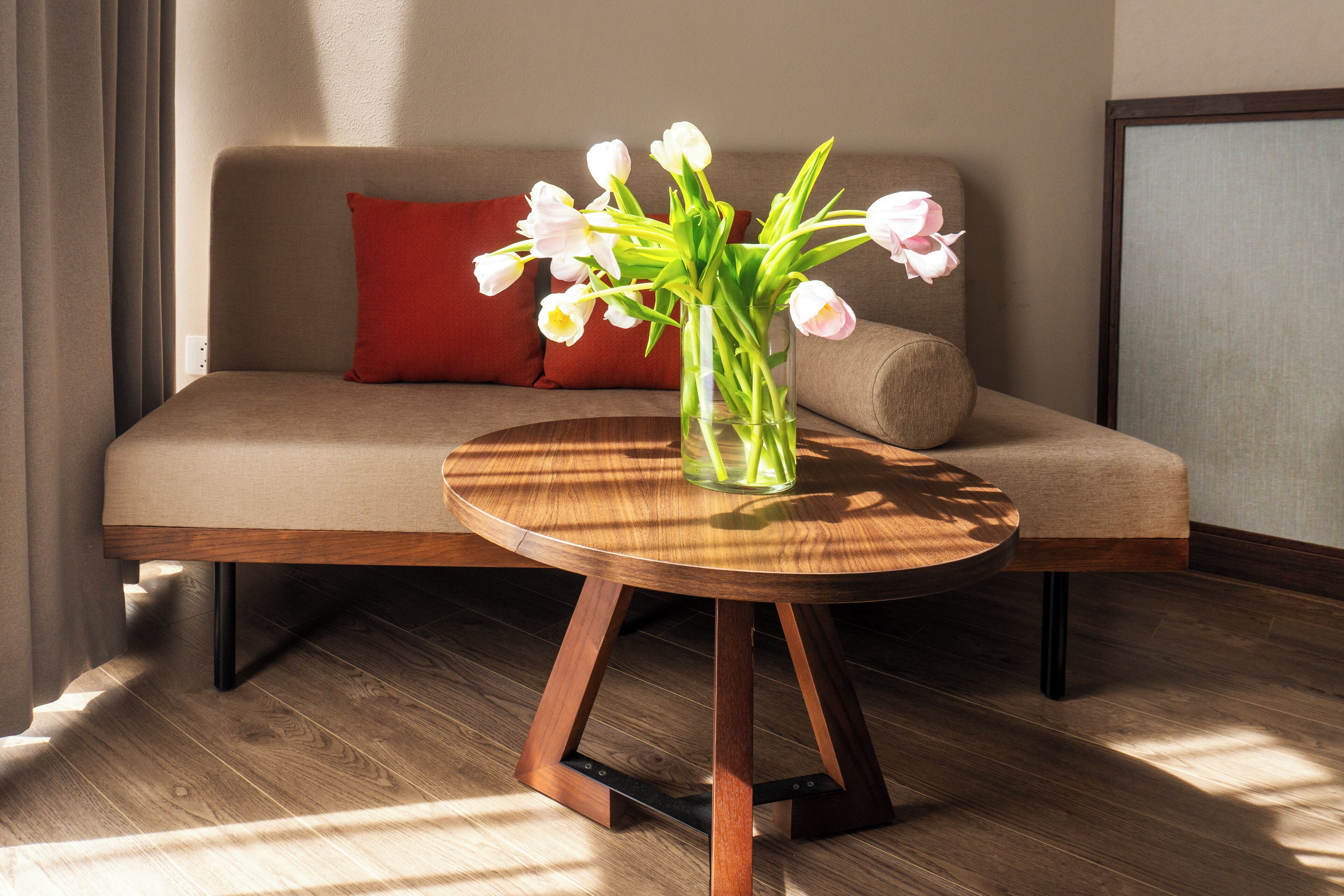 A modern living room with a round wooden table, a beige sofa with a red pillow, and a vase of white tulips.