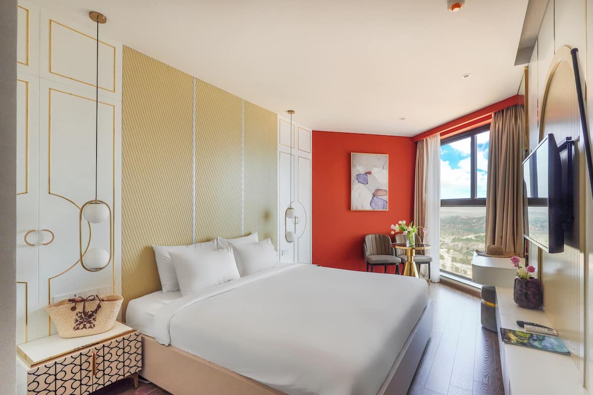 Una habitación de hotel moderna con una cama grande, un escritorio con silla, una mesita con un florero y una ventana con vistas.