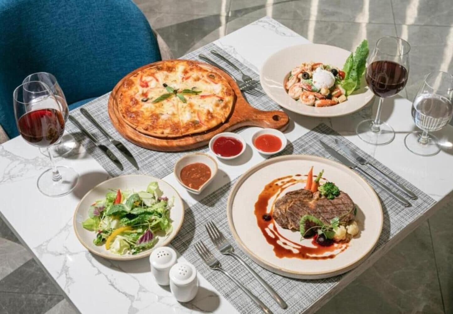 A table set with a pizza, pasta, steak, salad, and wine glasses.